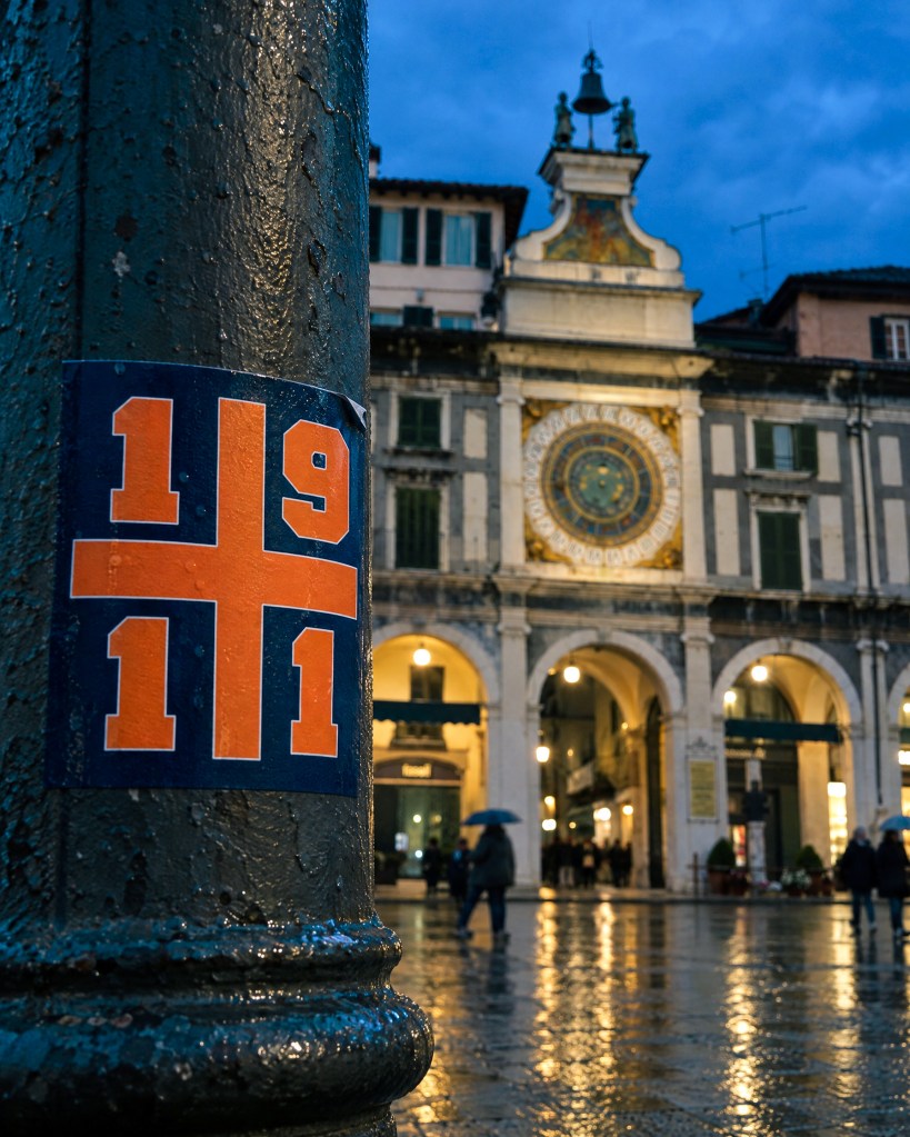 Nuovo adesivo 30 ottobre 2026: adesivo «1911» con la croce arancione su sfondo blu, attaccato a una colonna in piazza della Loggia a Brescia